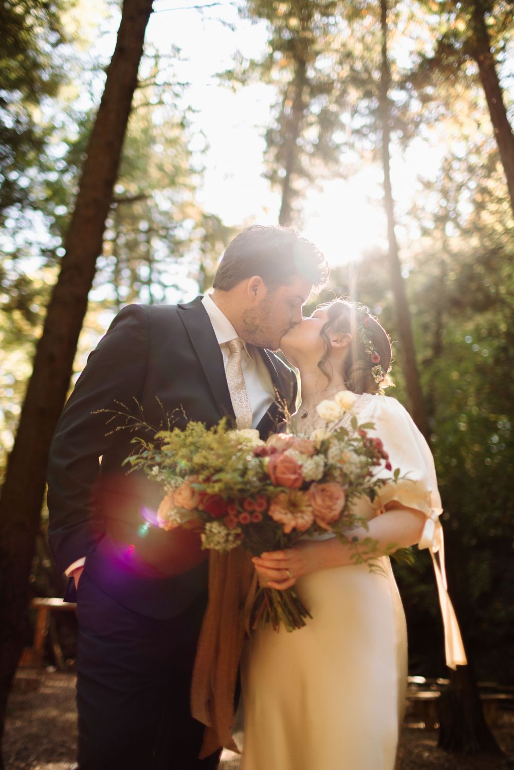 A romantic wedding moment captured in a sunlit forest with a beautiful bouquet of pink and white flowers.