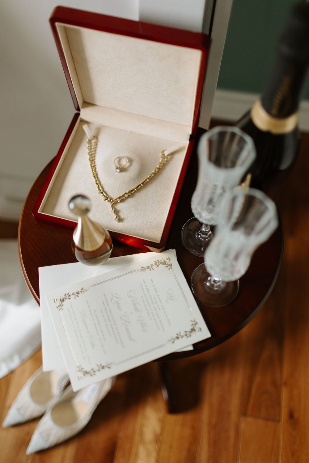 Wedding accessories including pearl jewelry and white shoes arranged on wooden floor with invitation card and gift boxes.