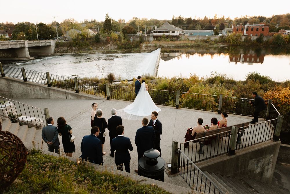 A wedding ceremony taking place on a scenic riverside terrace with guests gathered on stone steps during autumn.