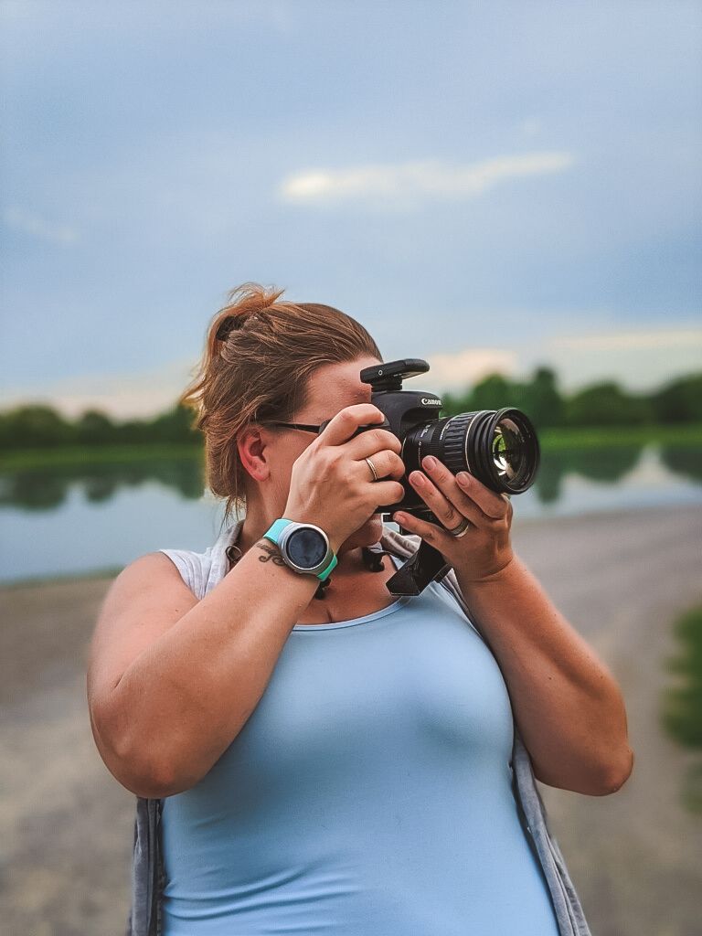 Photographer taking pictures outdoors by a lake wearing light blue tank top and smartwatch.