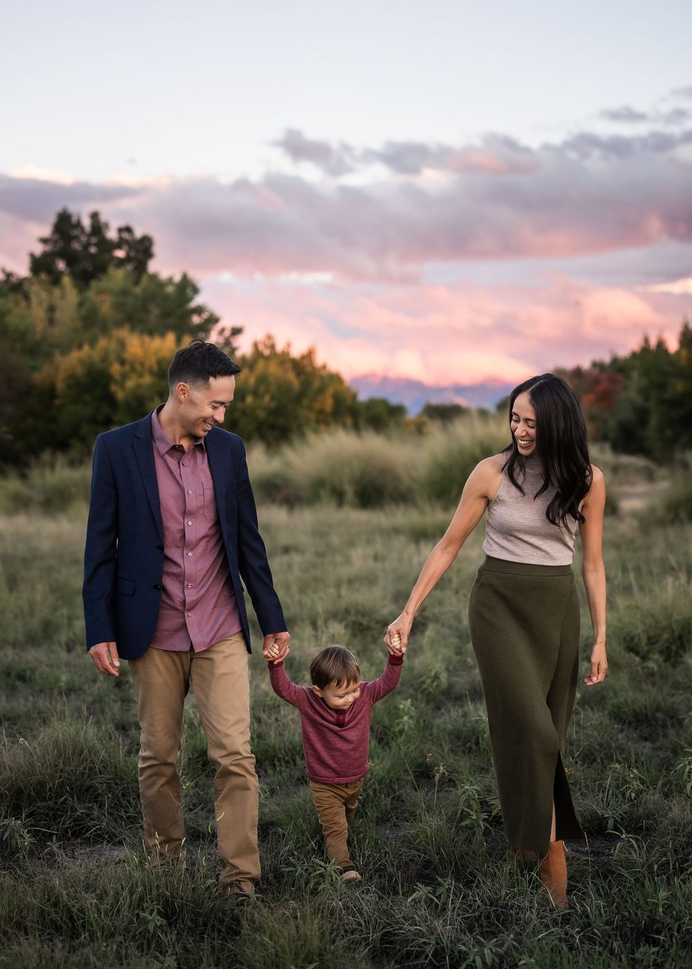 Family walking together in a grassy field at sunset with pink clouds and mountains in the background.