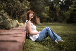 Person in white tank top and blue jeans sits on grass near brick wall with cactus plants in outdoor garden setting.