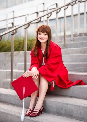 Graduate in bright red cap and gown sits on concrete steps holding diploma in hand while wearing burgundy flats.