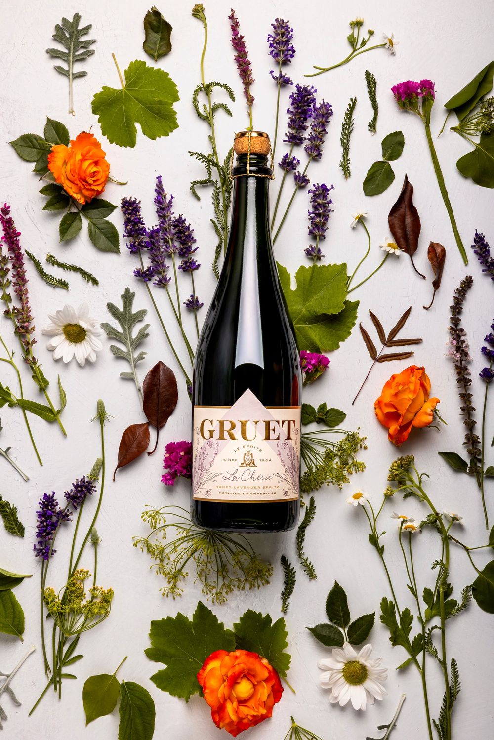 Gruet sparkling wine bottle surrounded by colorful flowers and herbs on white background.