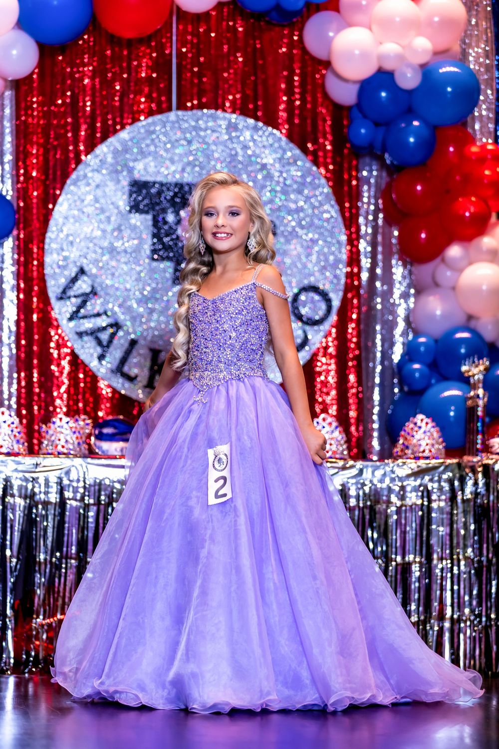 Pageant contestants in matching lavender ball gowns pose on stage with red curtain and balloon decorations.