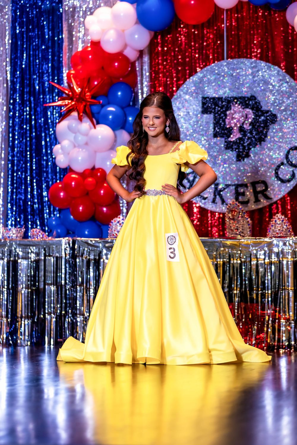 A contestant in a bright yellow ballgown poses on stage with red, white and blue balloon decorations and sparkly backdrop.