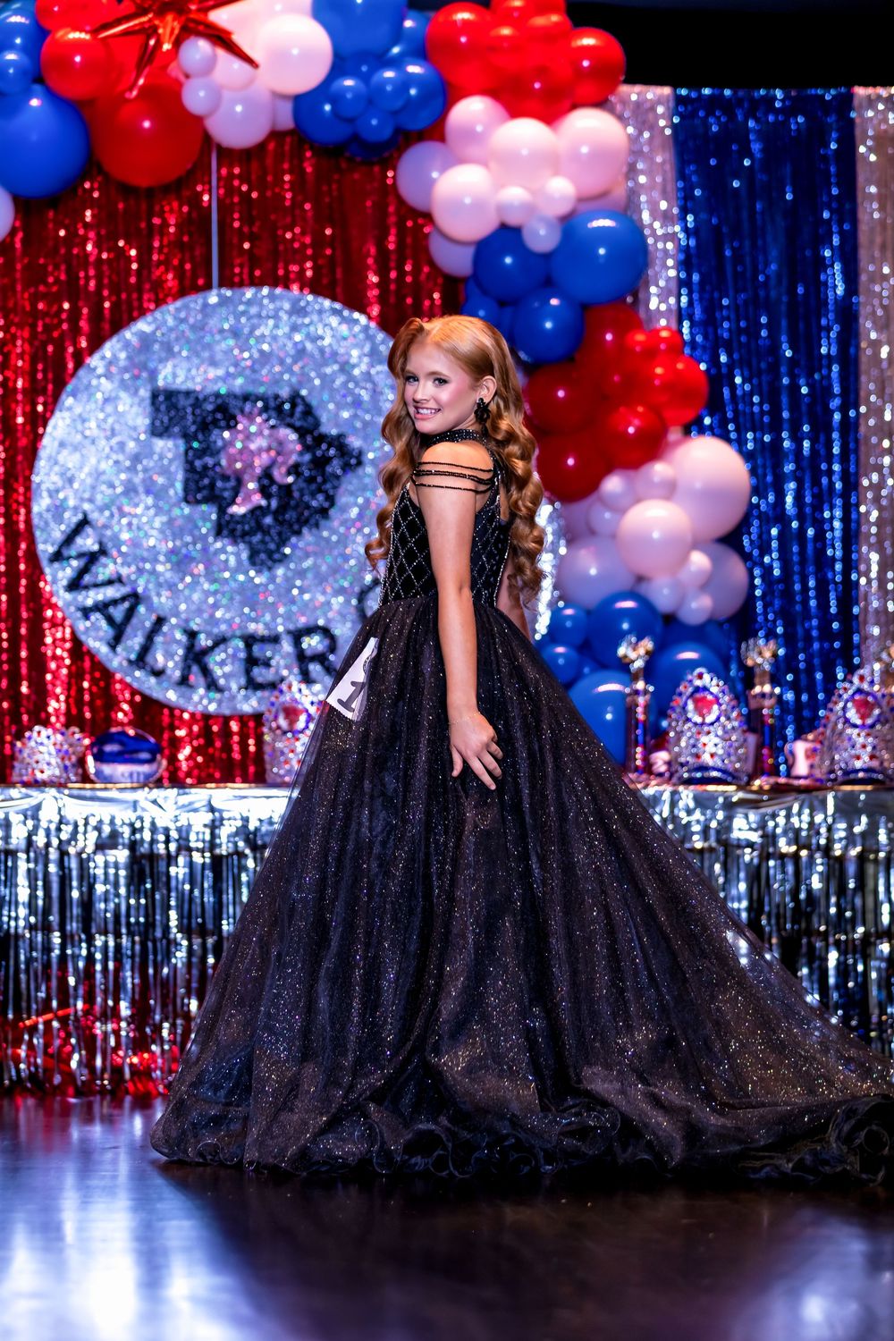 Sparkling black ball gown on stage with red, white and blue balloon decor against a shimmering curtain backdrop.