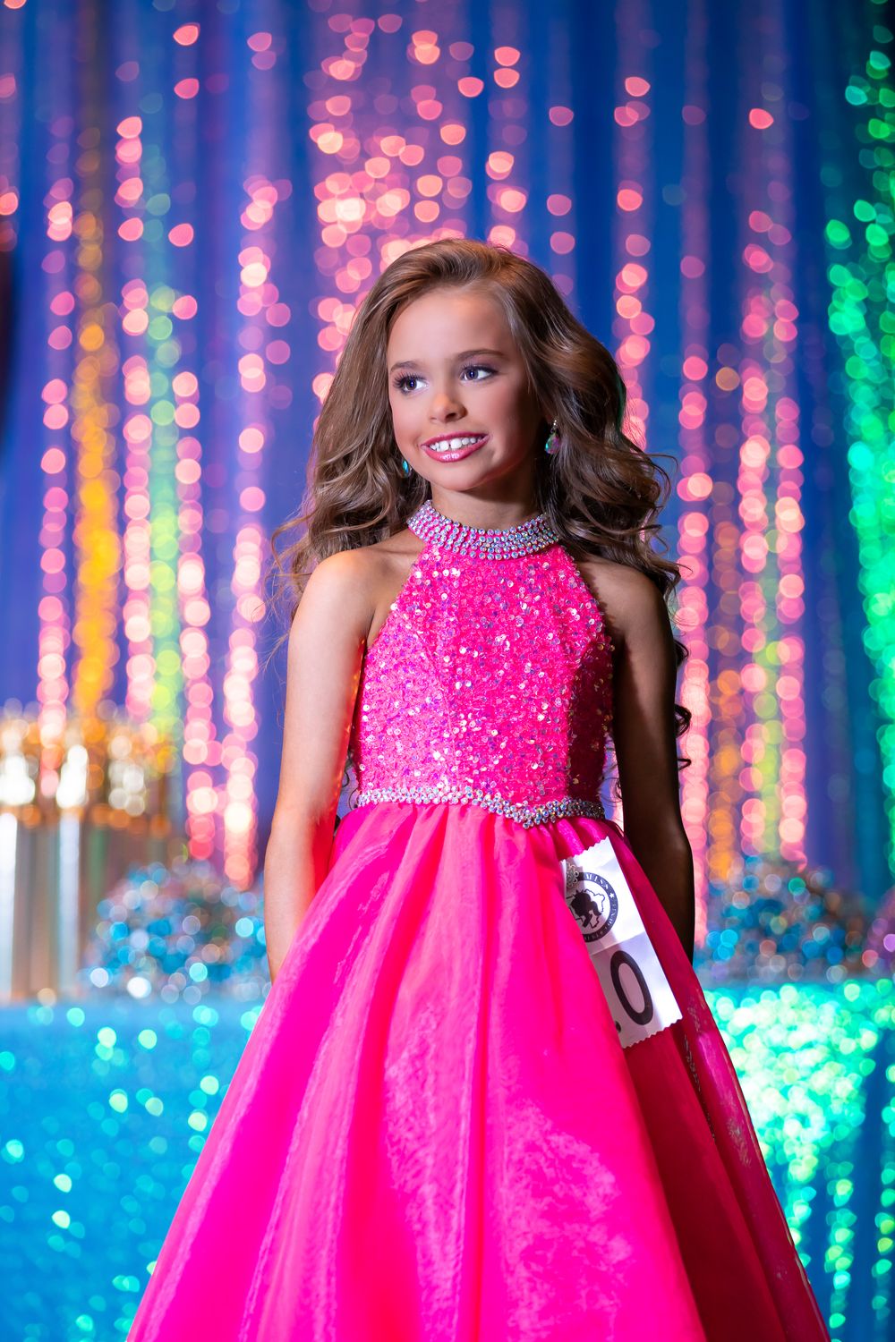 Pageant performer in dazzling pink gown stands against colorful light curtain backdrop.