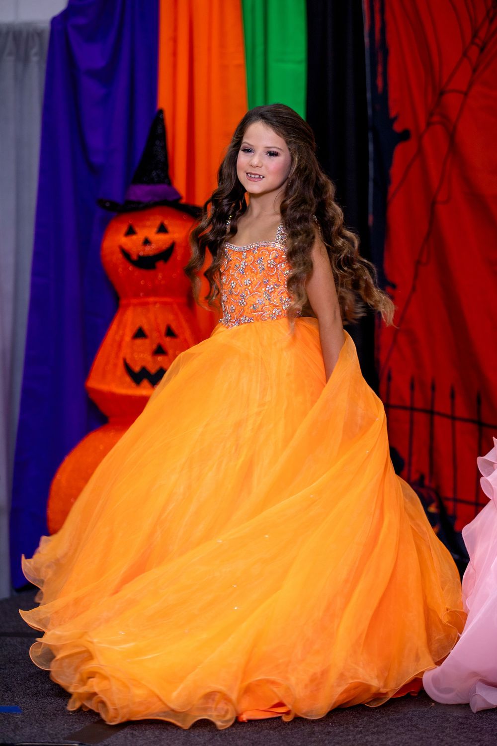 A bright orange tulle ball gown with ruffled layers stands out against a colorful backdrop of purple and red drapes.