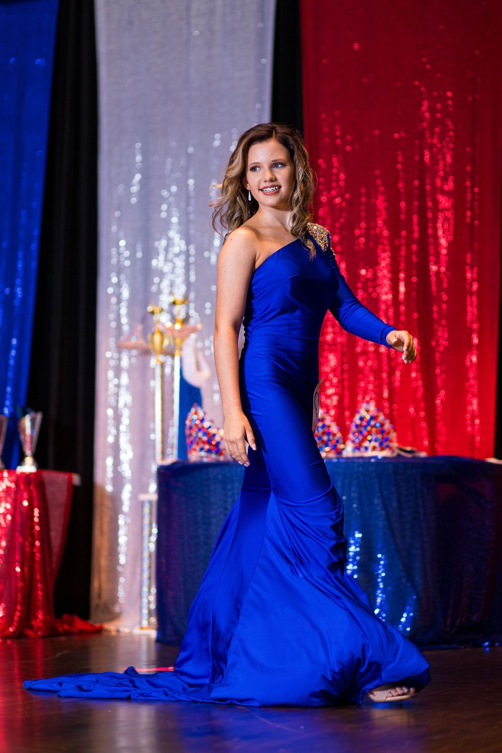 Elegant royal blue one-shoulder mermaid gown with dramatic train on stage with sparkling backdrop.