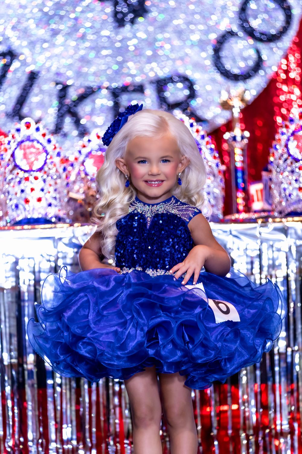 Sparkly blue pageant dress with sequins and ruffles against a patriotic red white and blue background with glitter.