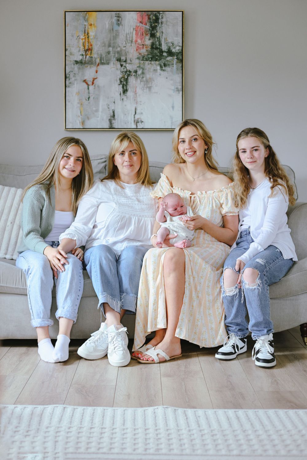 Four people sit together on a white couch beneath modern artwork, holding an infant in a peaceful home setting.