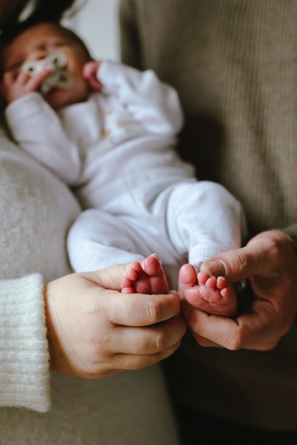 Close-up of small baby feet being held by gentle hands while infant rests in white clothing.