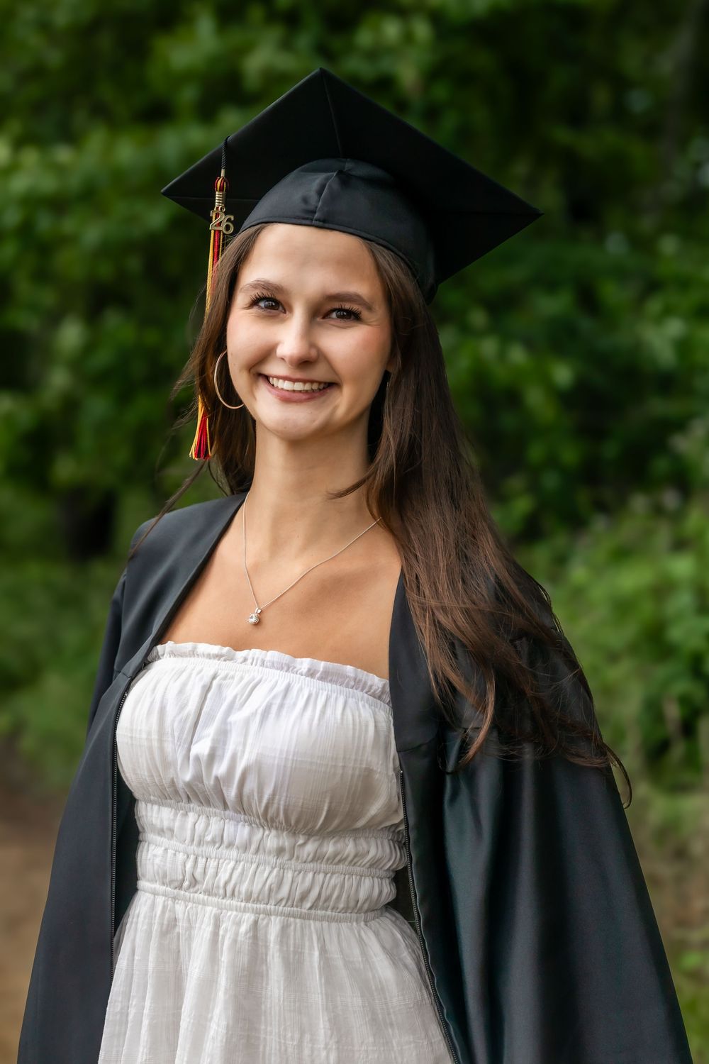 Smiling young woman in black graduation cap and gown over white dress, posing outdoors with green trees in background.