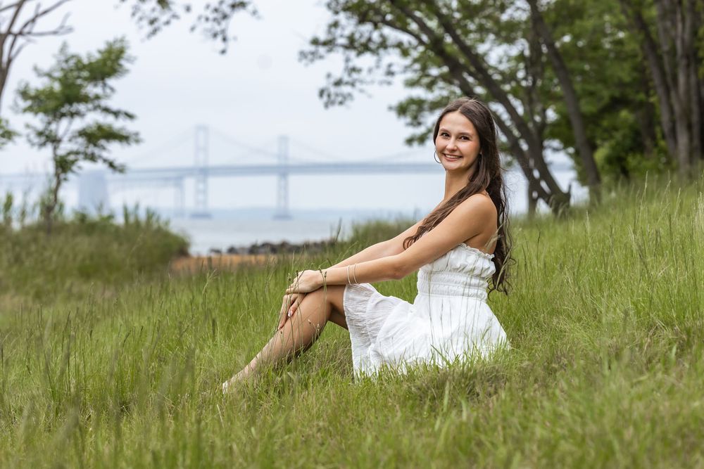 Young woman in white dress sitting on lush green grass with a scenic bridge and water view in the background.