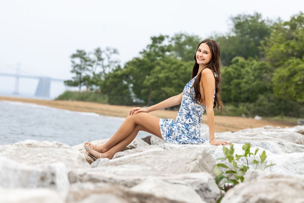 Young woman in blue floral dress sitting on rocky shoreline with trees and bridge in background.
