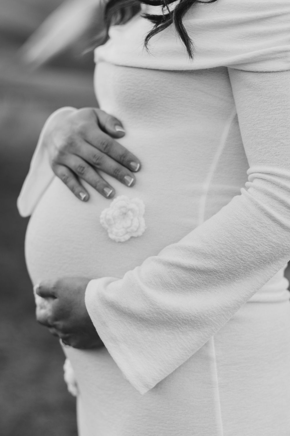 Black and white photo of pregnant woman in white dress cradling baby bump with small flower decoration.