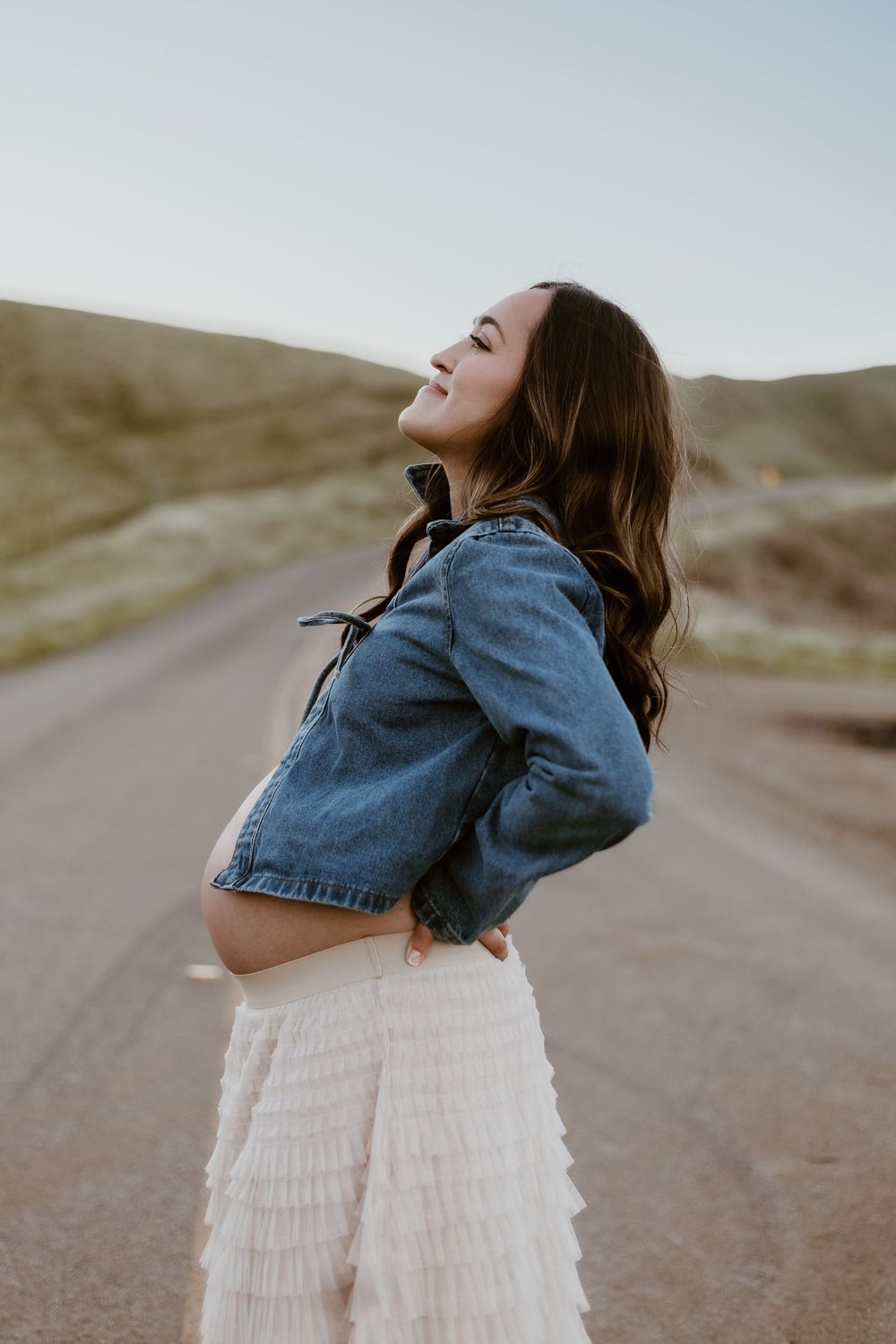 Pregnant woman in denim jacket and white ruffled skirt poses on a road, smiling with head tilted back.