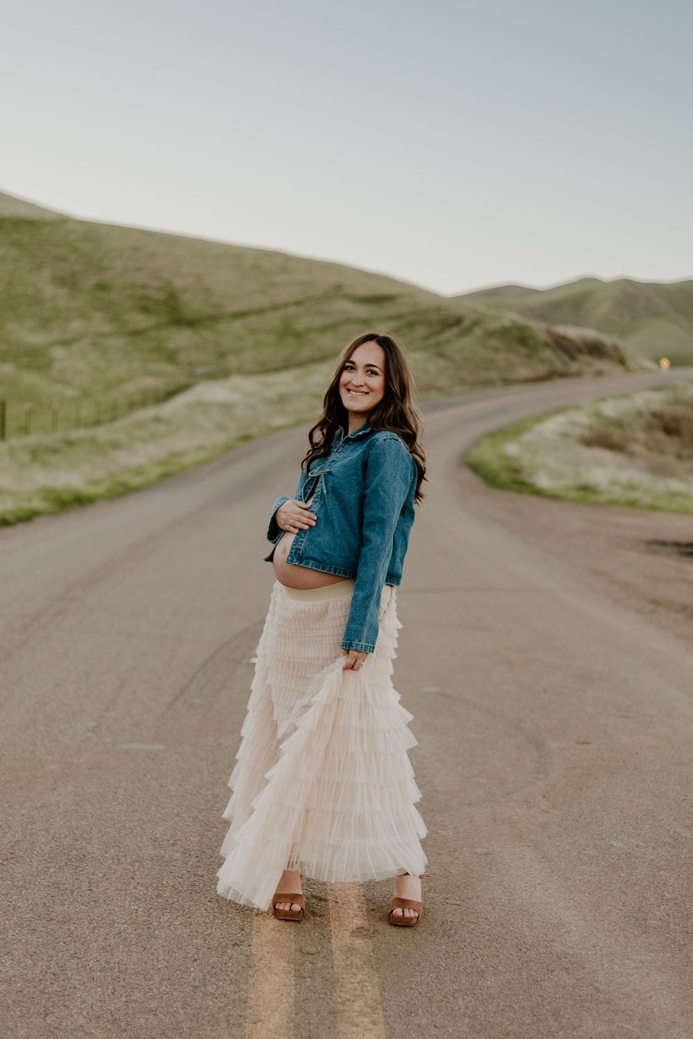 Pregnant woman in denim jacket and white ruffled maxi skirt standing on a scenic rural road with green hills.