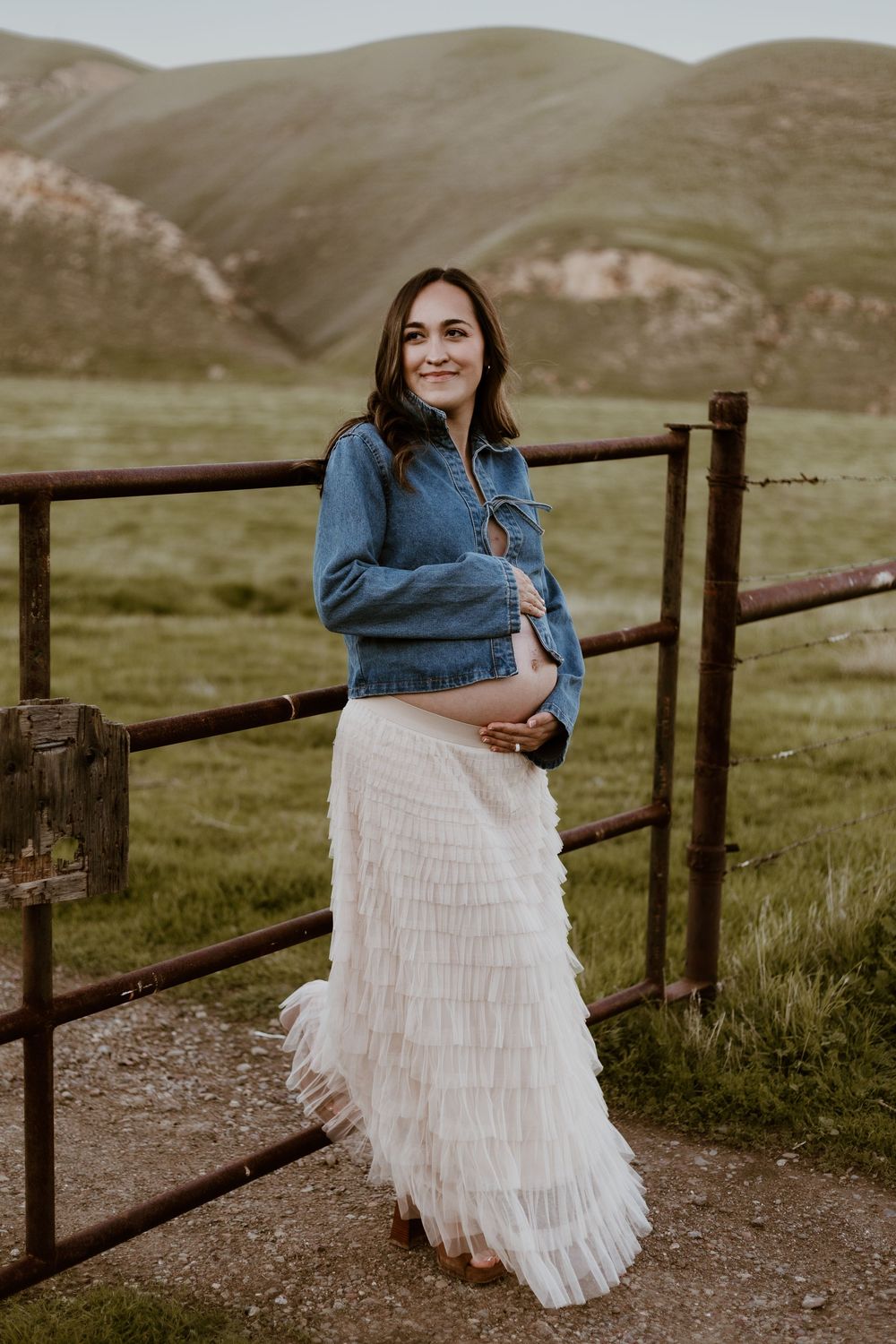 Pregnant woman in denim jacket and white ruffled skirt leaning on rustic fence with green hills backdrop.