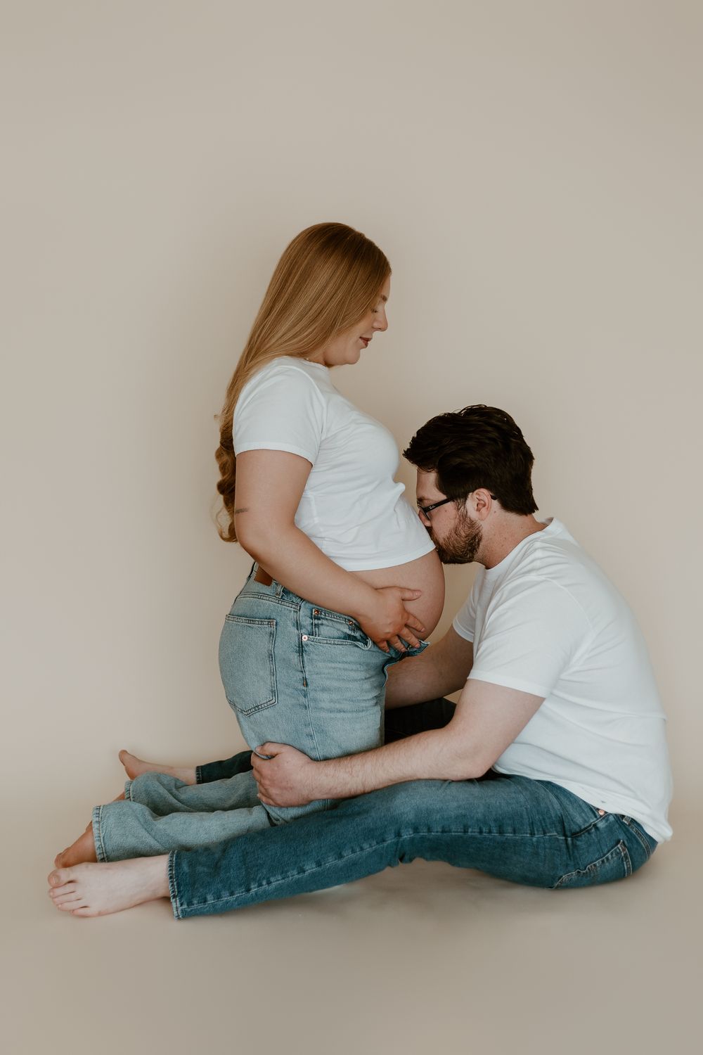 A man sitting cross-legged kisses a pregnant woman's belly during a maternity photo shoot in matching white tees.