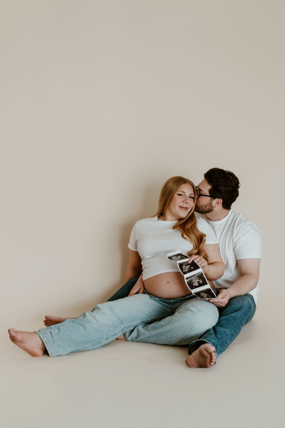 Pregnant couple sitting on floor holding ultrasound photos, woman in white crop top and jeans smiling at partner.