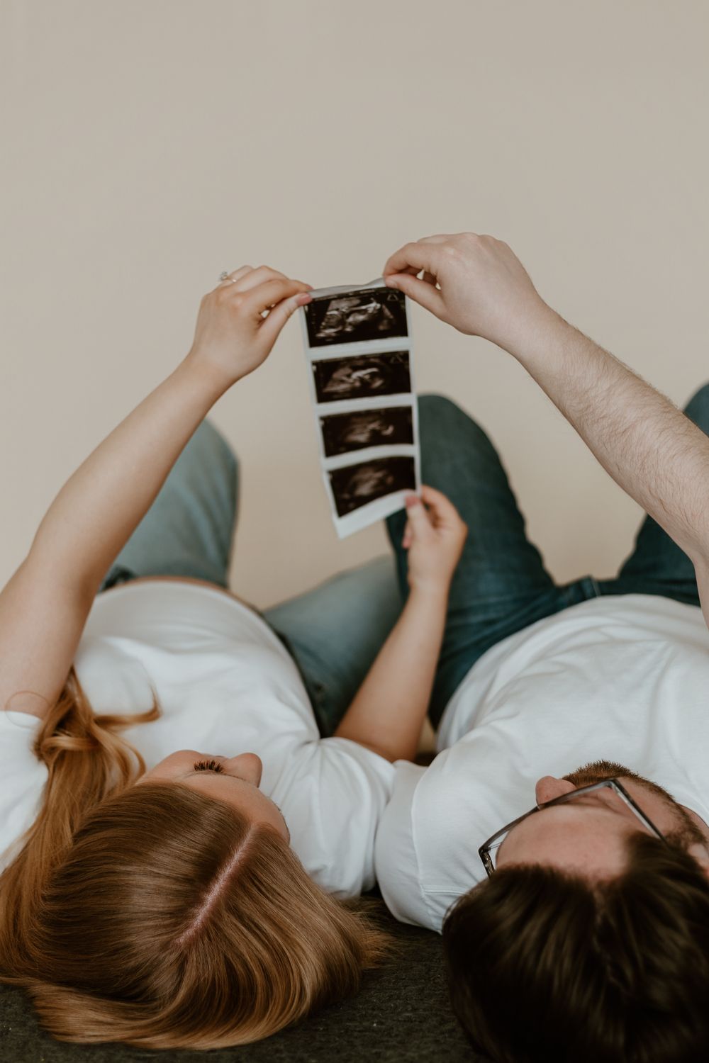 Couple lying down together holding ultrasound sonogram photos, expecting parents sharing a joyful pregnancy announcement moment.