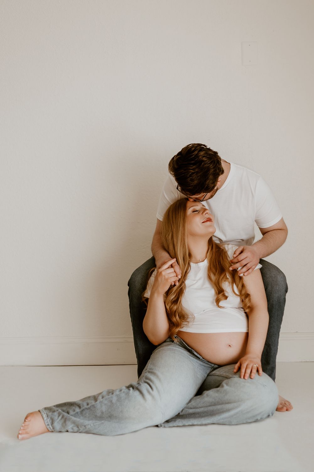 Pregnant woman sitting on chair with partner standing behind her, both in casual white tops and gray pants.