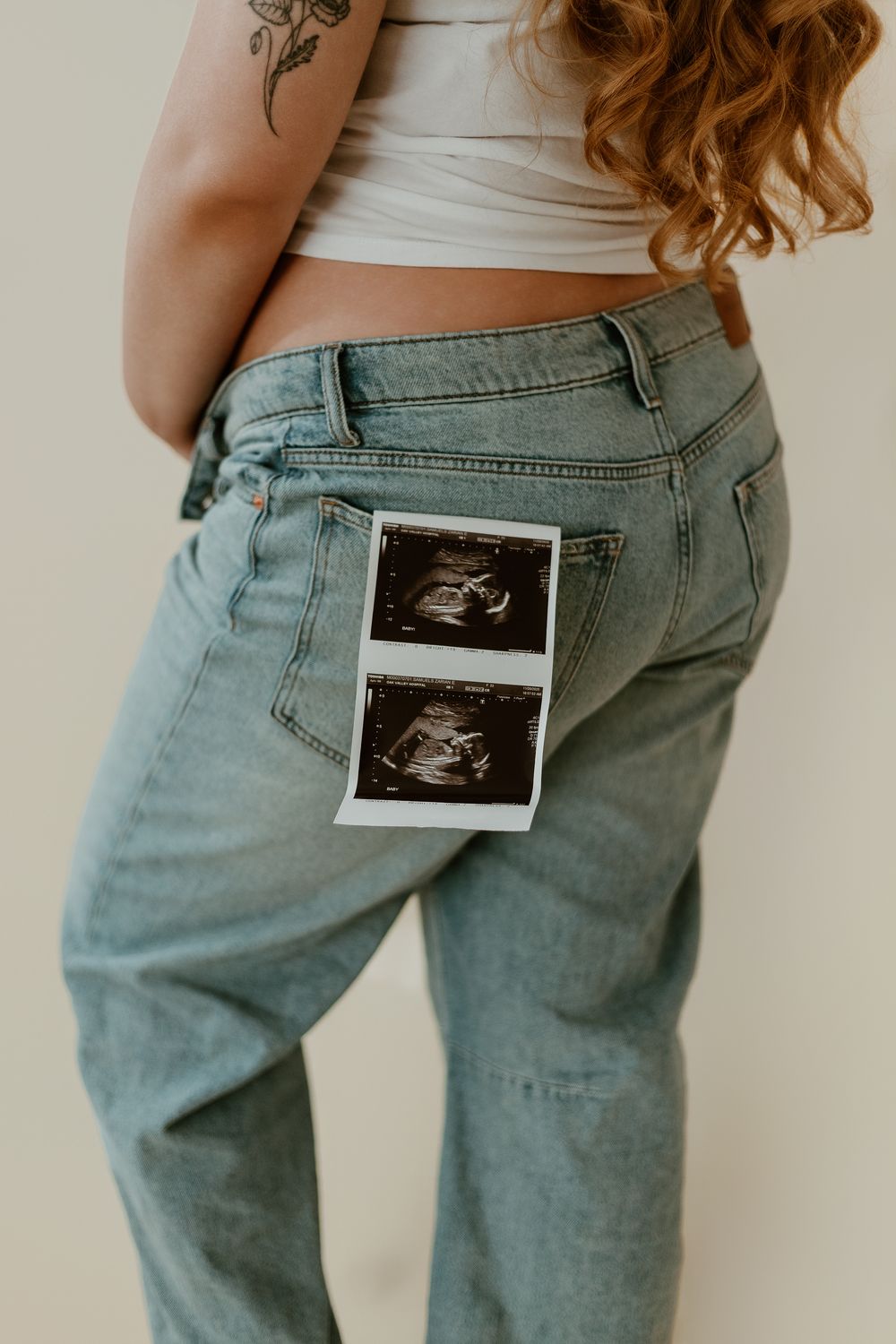 Pregnant woman in light wash jeans holding ultrasound photos in back pocket, wearing white crop top with floral tattoo.