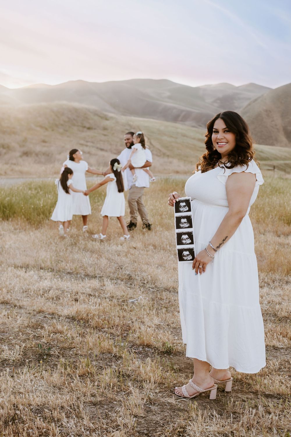 A family in white dresses poses in a golden grassy field with mountains at sunset while holding ultrasound photos.