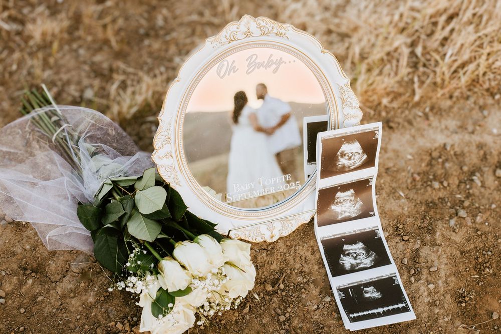 A vintage white frame with photos and ultrasound images beside white roses and eucalyptus on a rustic backdrop.