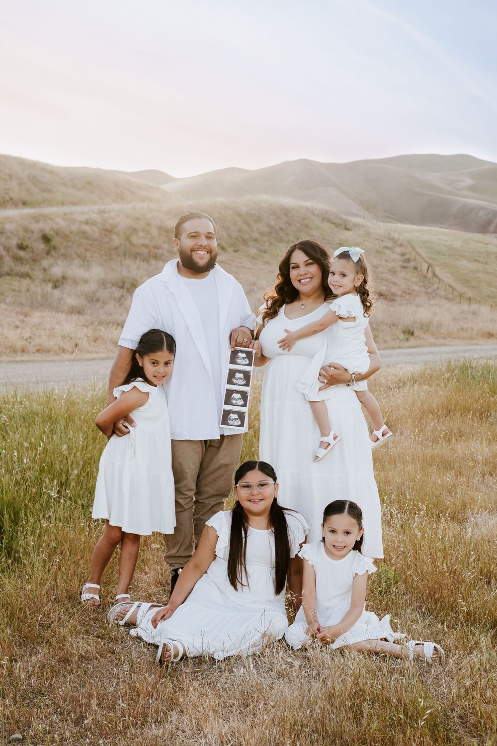 Family wearing coordinated white outfits poses together in grassy field with rolling hills at sunset.