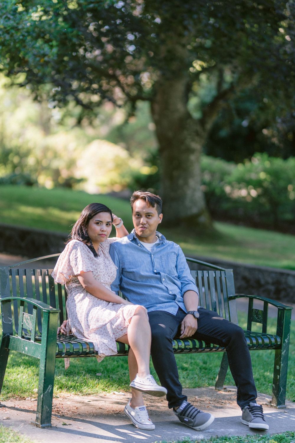 A couple sits together on a green park bench surrounded by lush garden scenery.