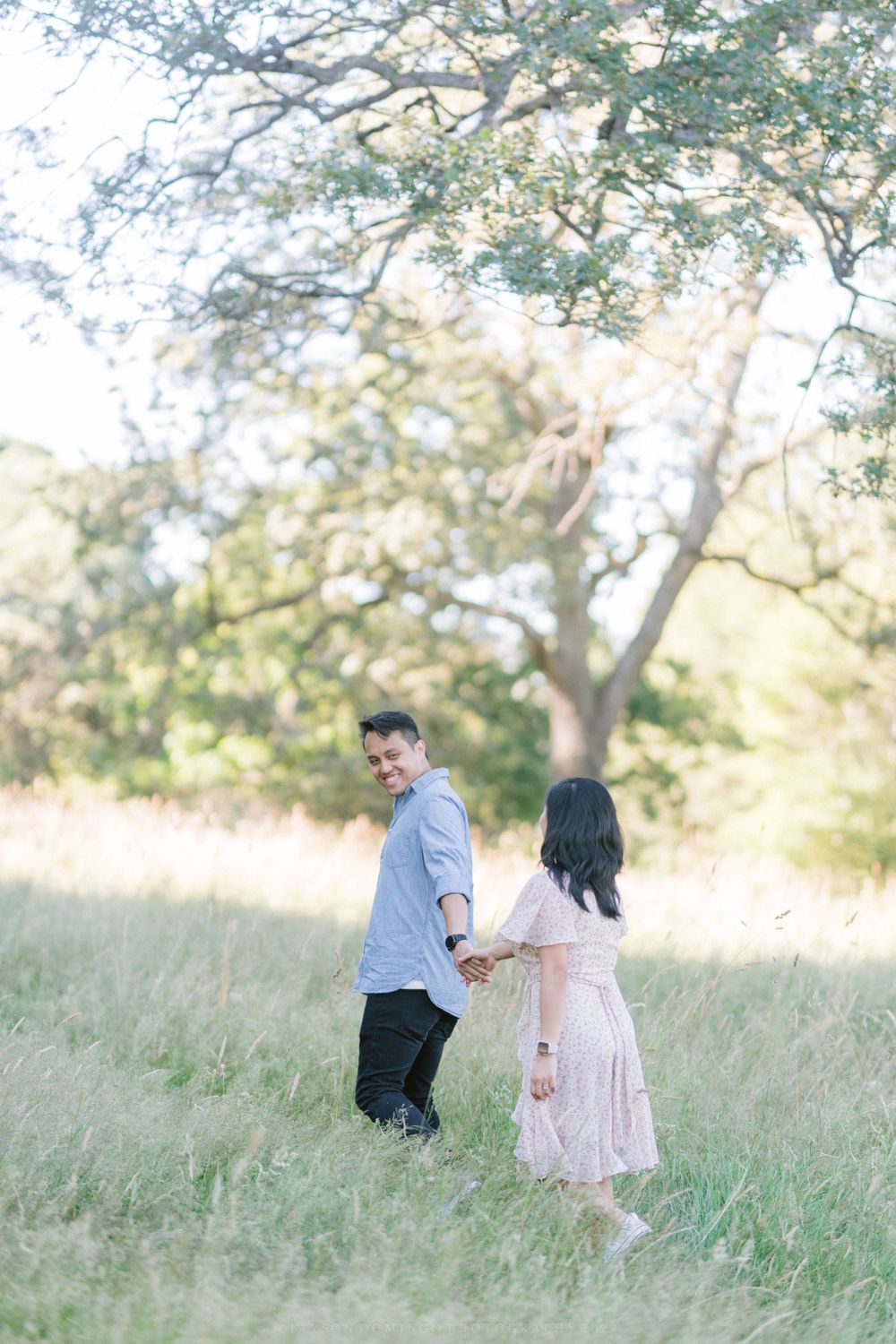 A romantic couple holds hands while walking through tall grass in a sunny meadow.