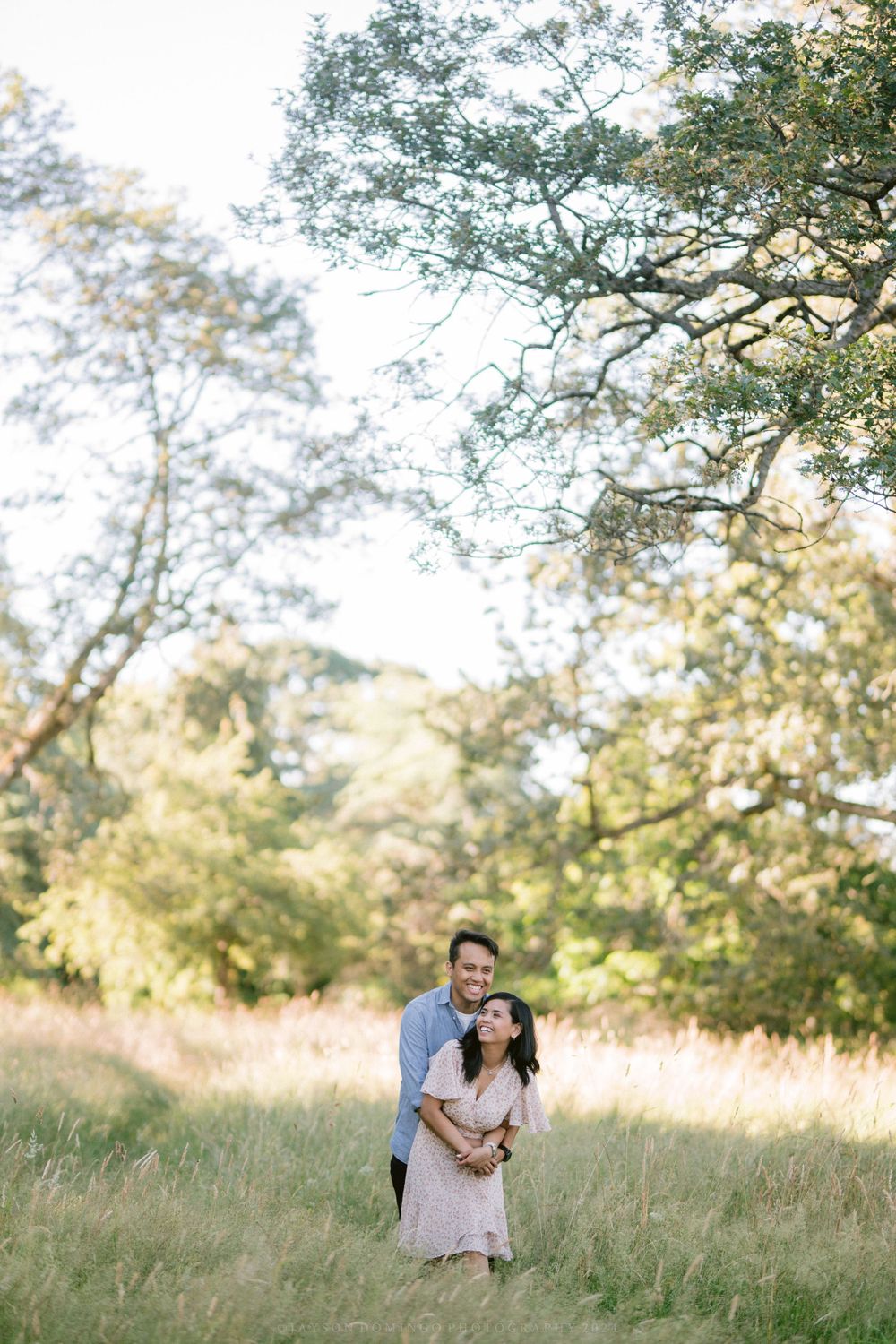 A loving embrace captured in a sun-dappled meadow surrounded by leafy trees.