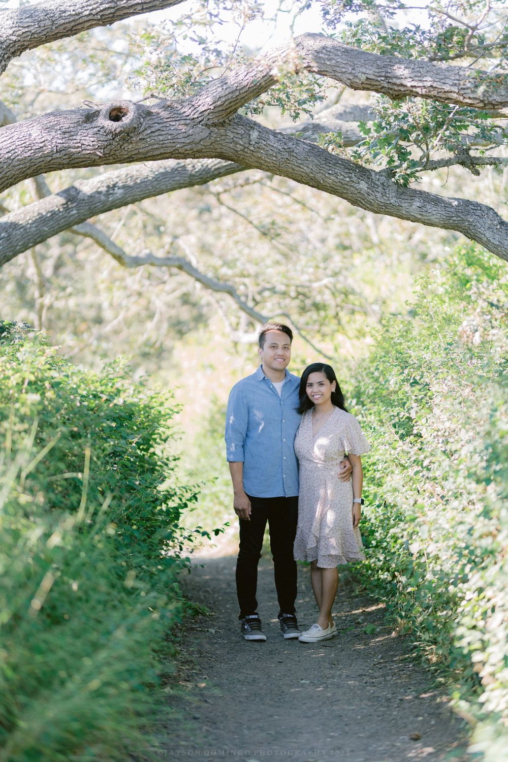 A couple stands together on a nature trail beneath an arching tree branch.