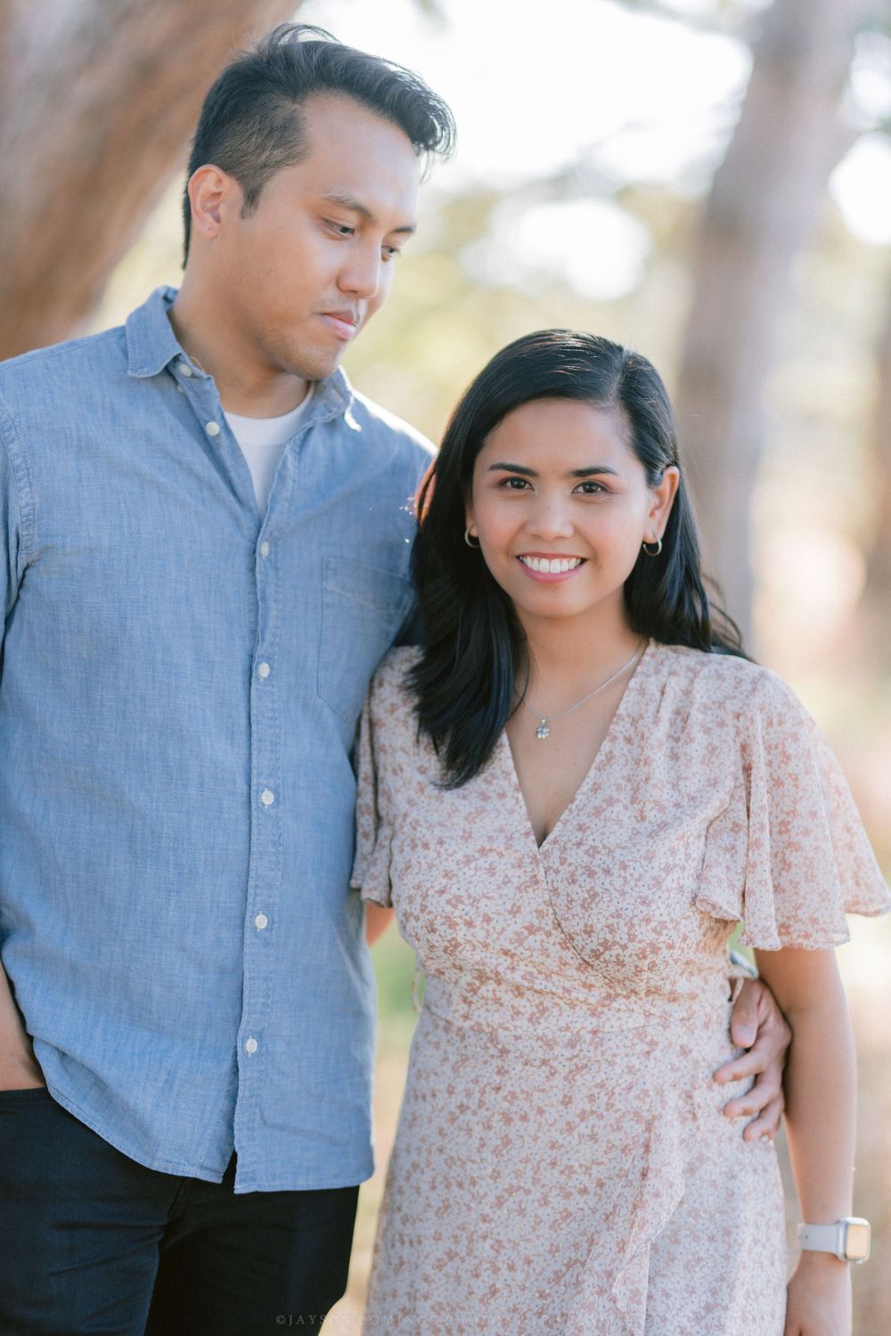 A couple in coordinating blue and pink outfits pose together in a sunlit outdoor setting.