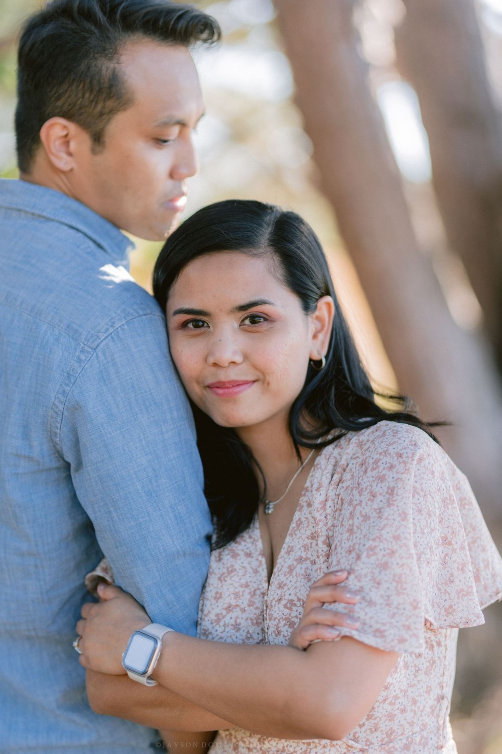 A romantic couple embraces while standing among trees in soft natural lighting.