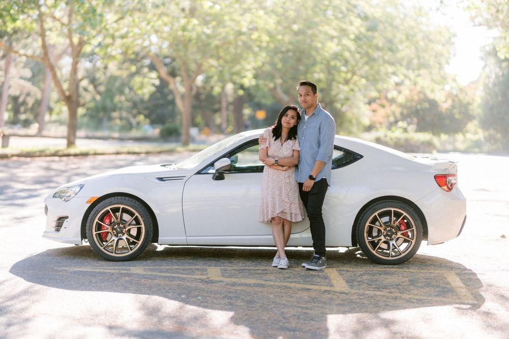 A couple poses beside a white sports car in a sunny park setting.