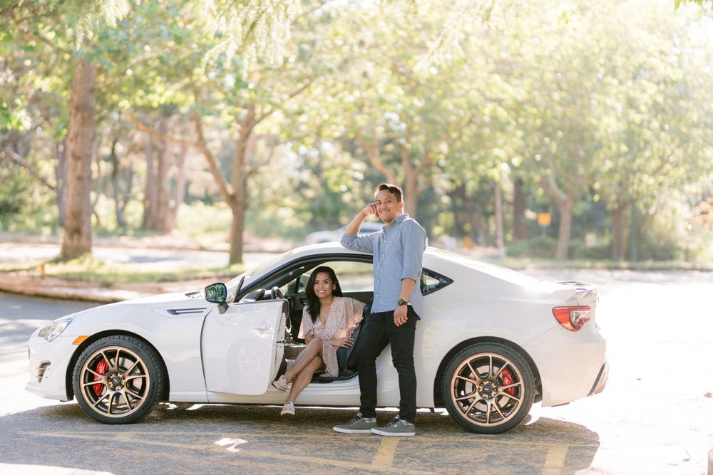 A couple stands by their white sports car as one person sits in the driver's seat.