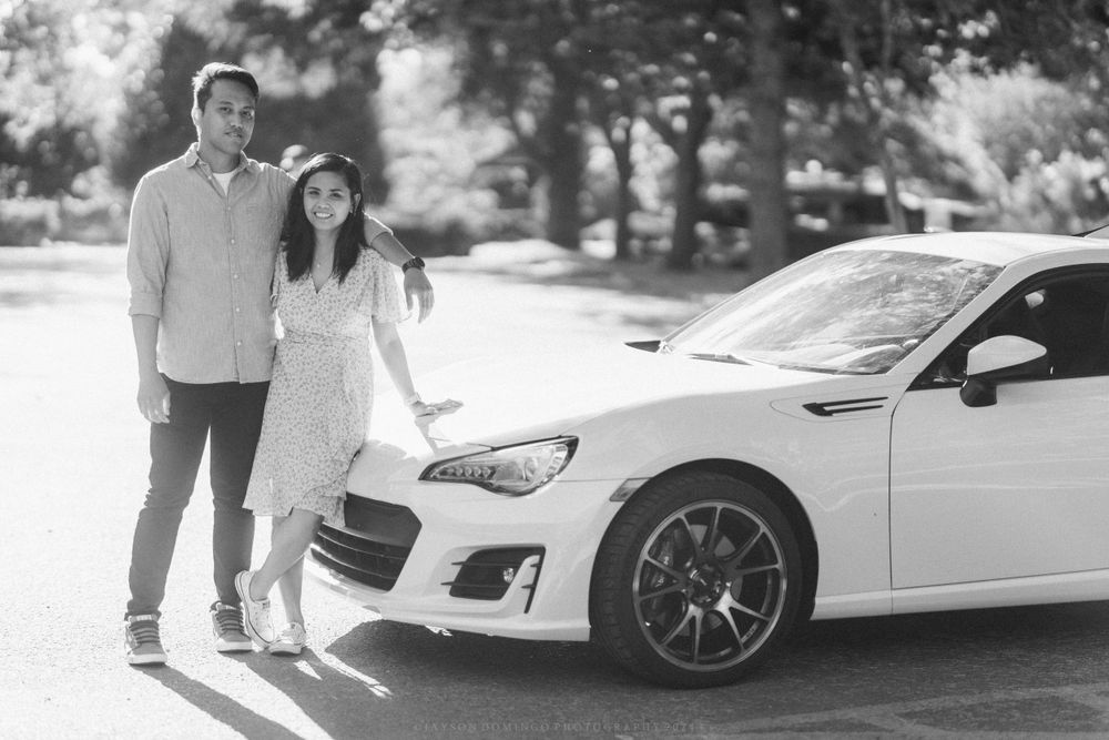 A couple poses next to their white sports car in a black and white photograph.