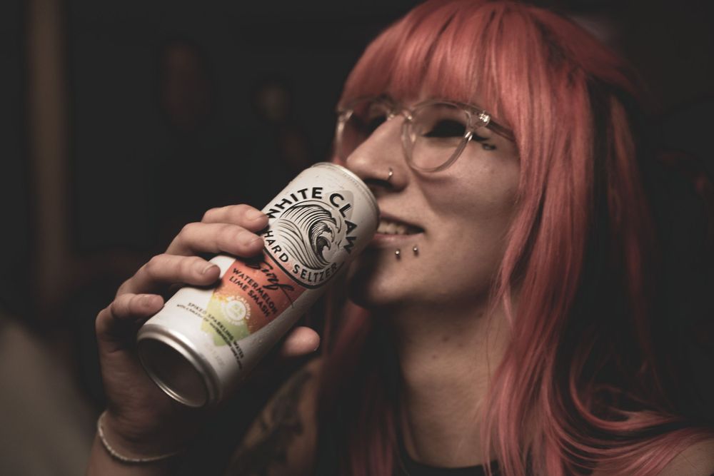 Person with pink hair drinking from various beverage cups in dark moody lighting.