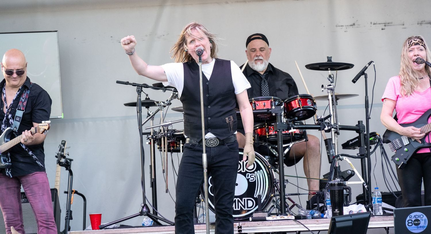 Band performing on outdoor stage with guitars and drums under white tent.