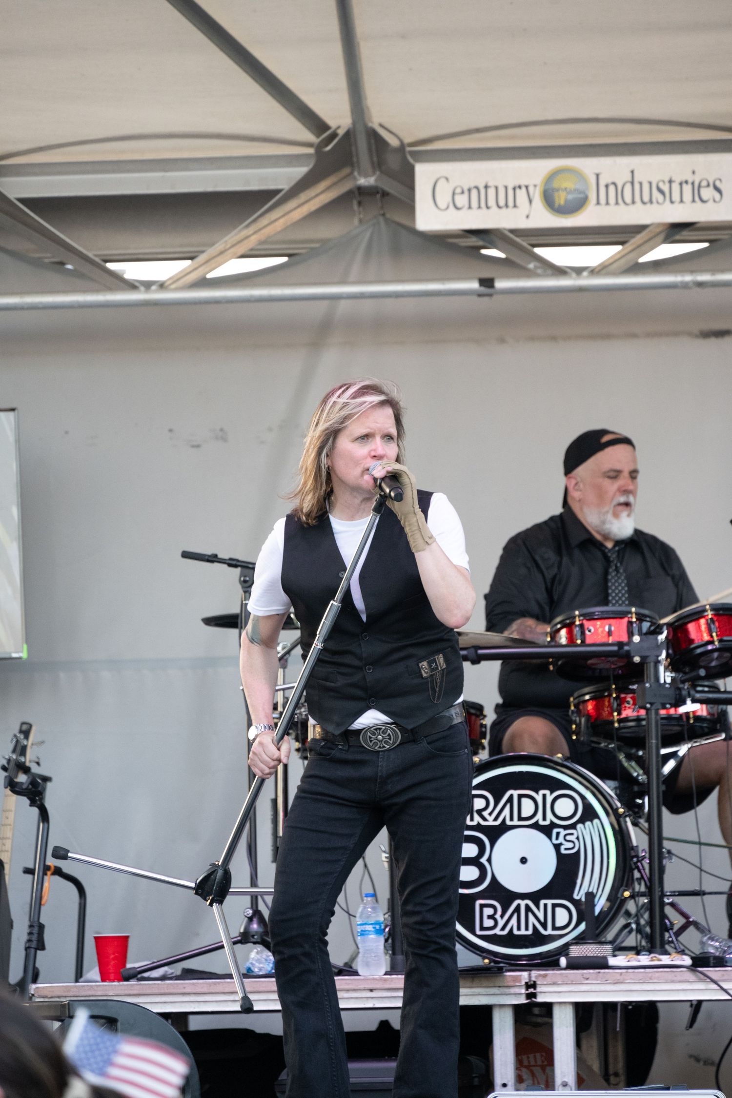 Band member performing on stage under Century Industries sign with drums in background.