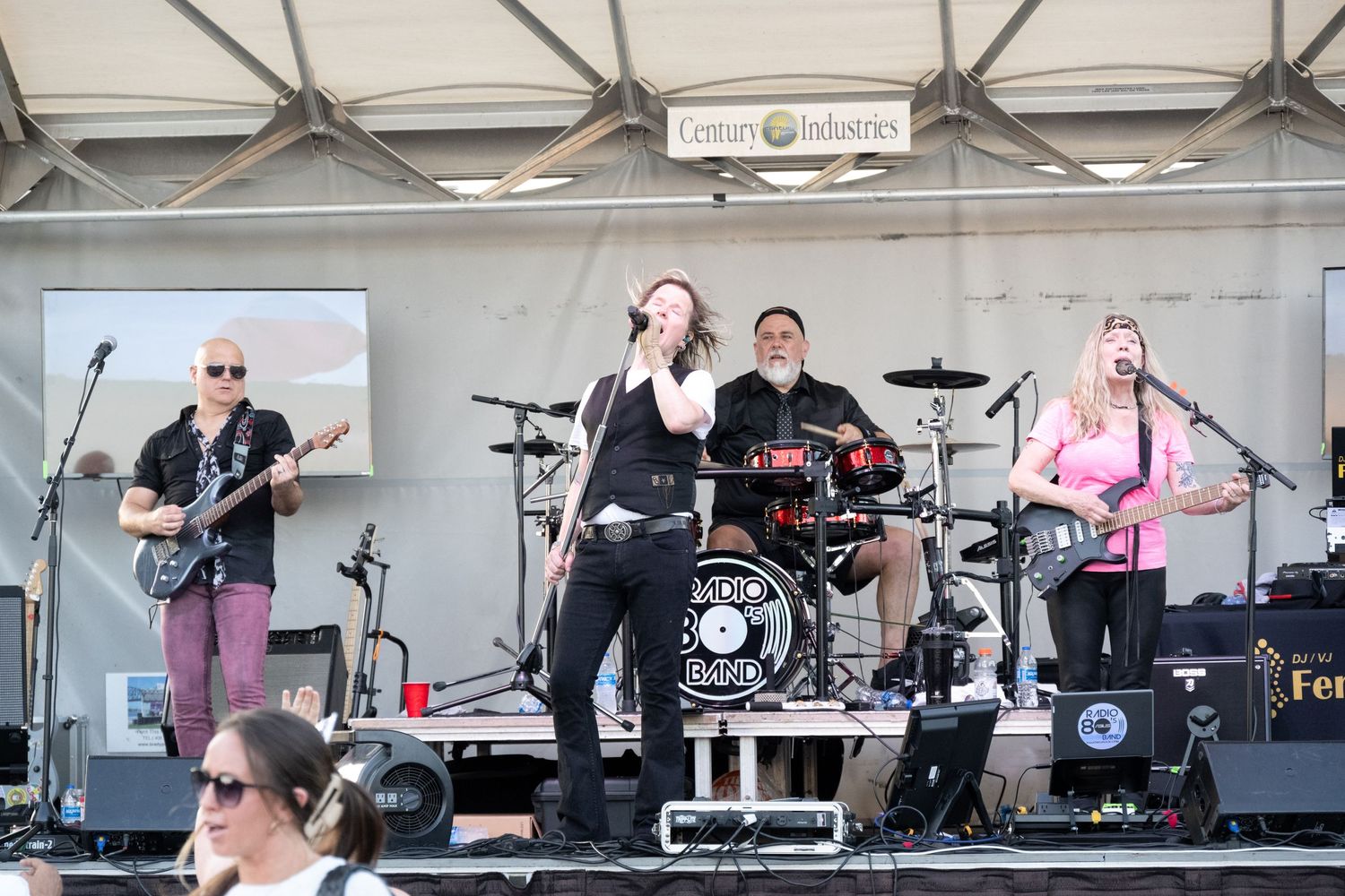 Live band performs on outdoor stage with guitar, drums and vocals during daytime concert performance.