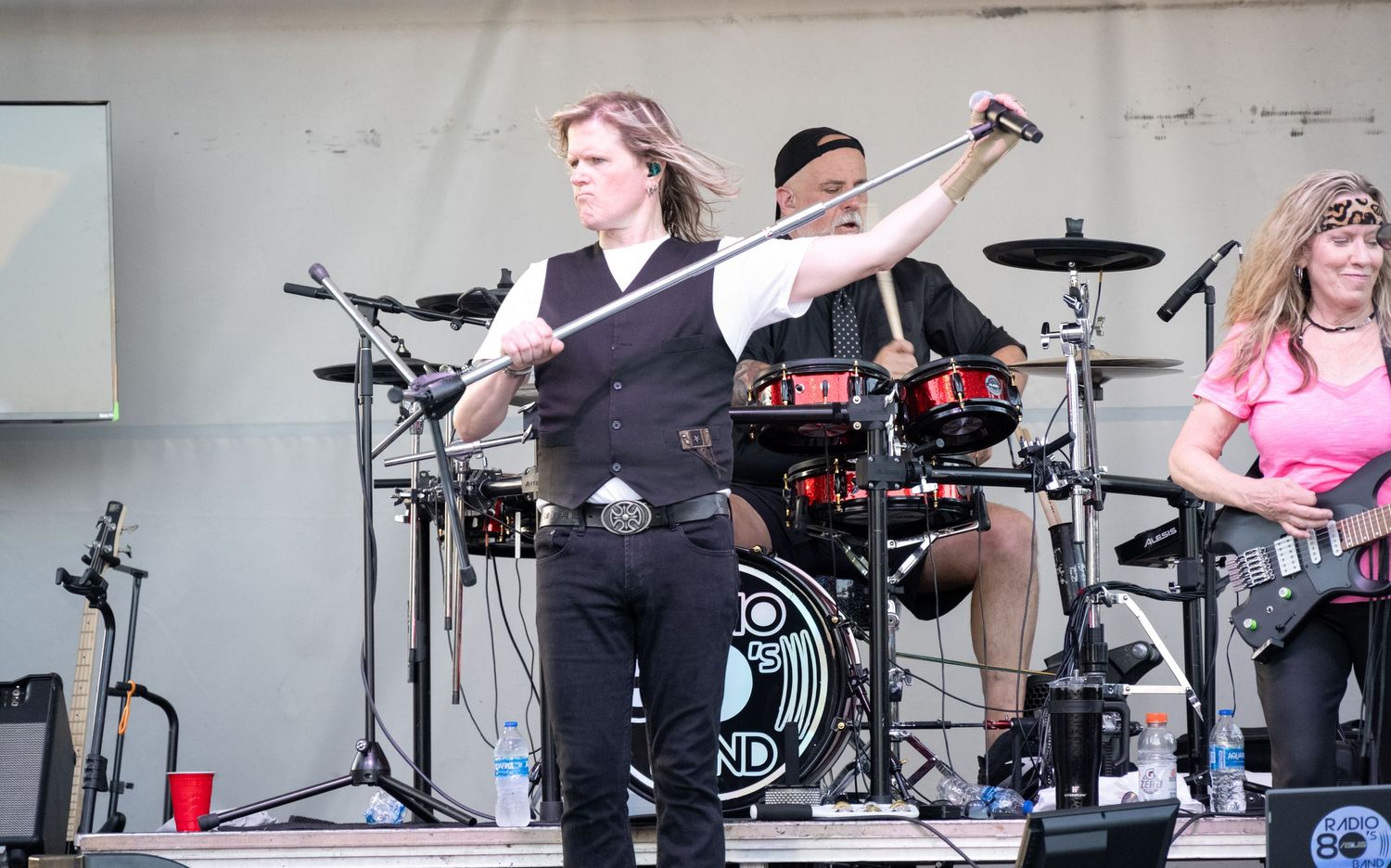 Musicians perform on stage with drum kit and various instruments during outdoor show.