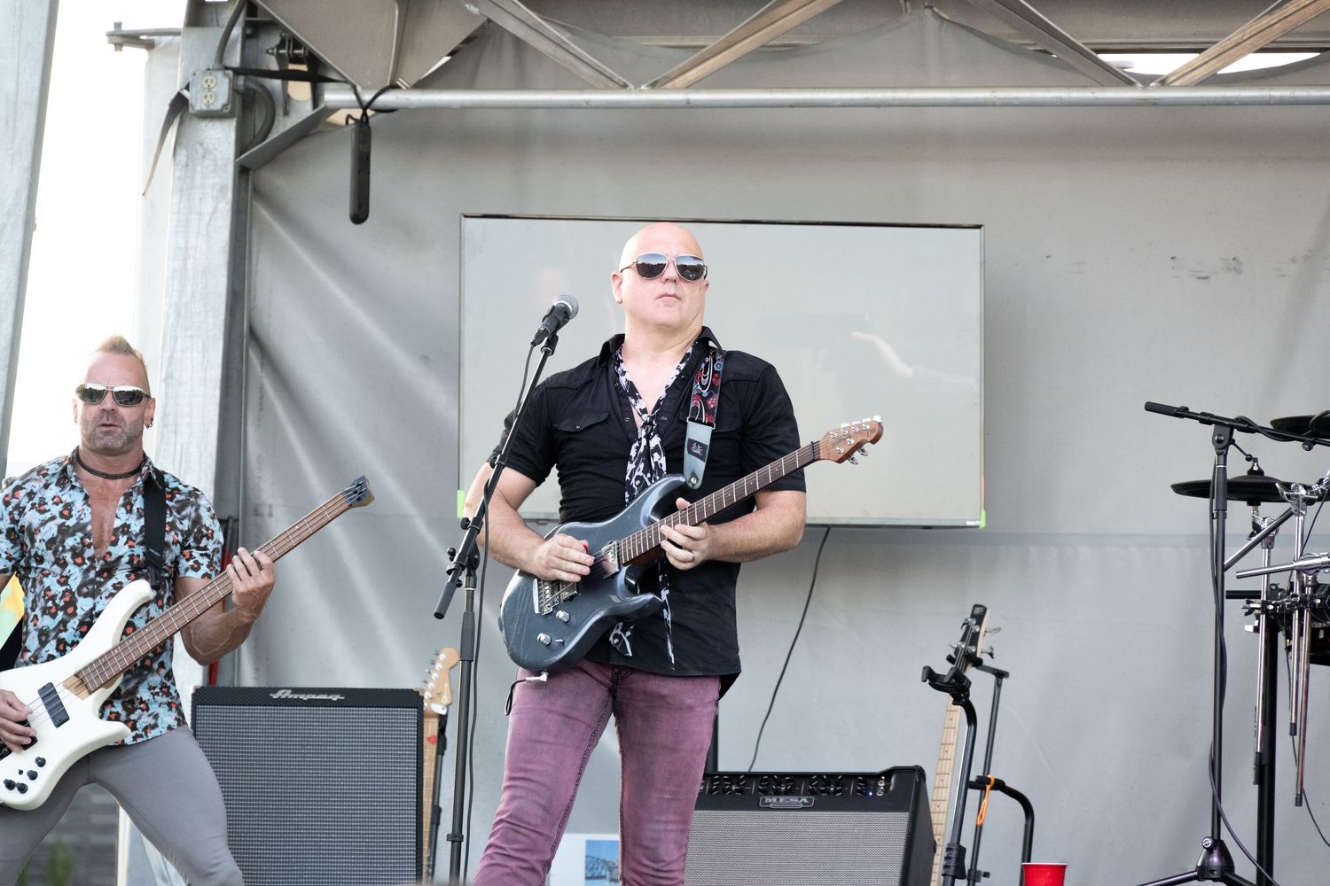 Musicians playing guitars perform on outdoor stage during daytime concert event.