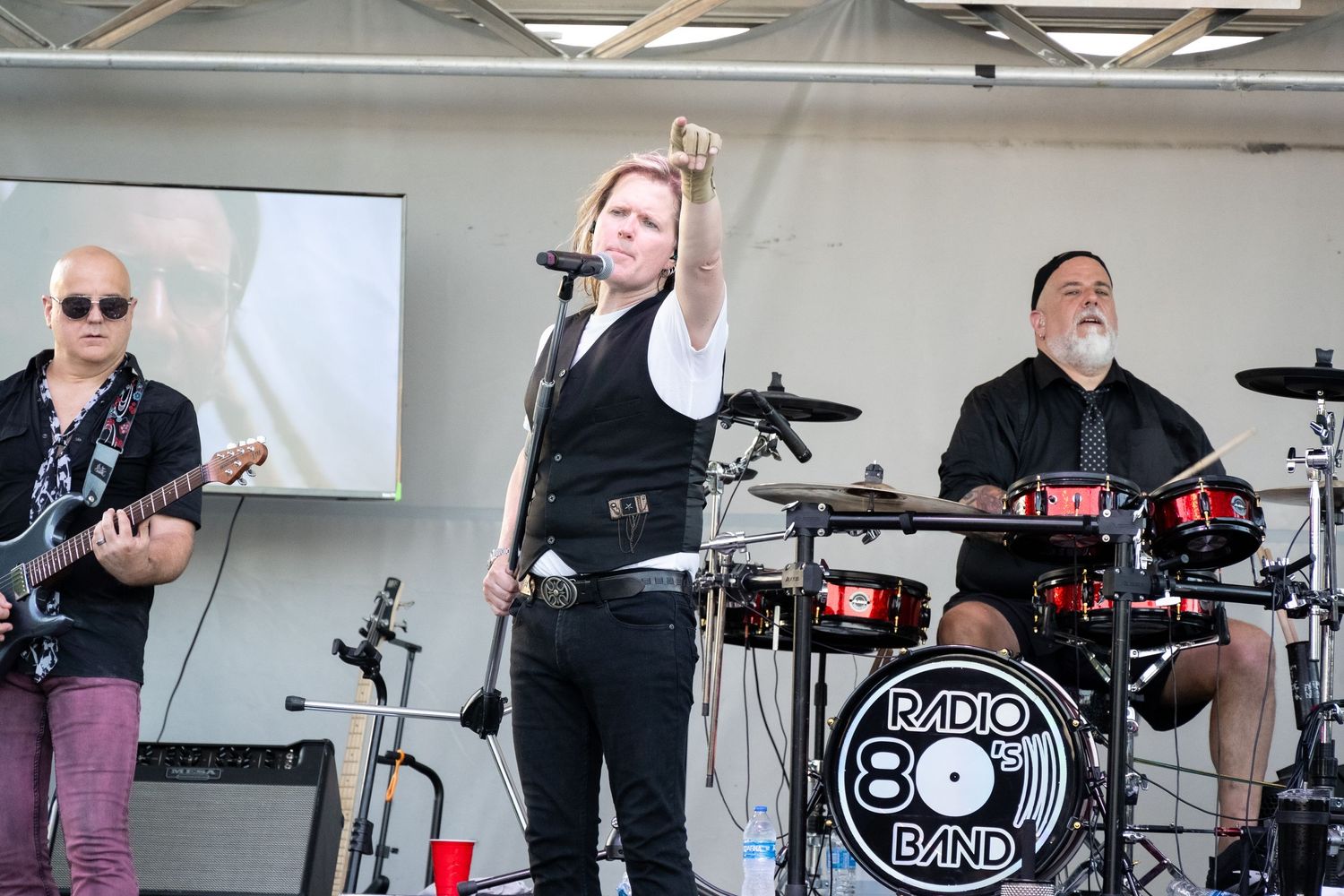 Rock band performs energetically on stage with Radio 80s Band drum kit visible.