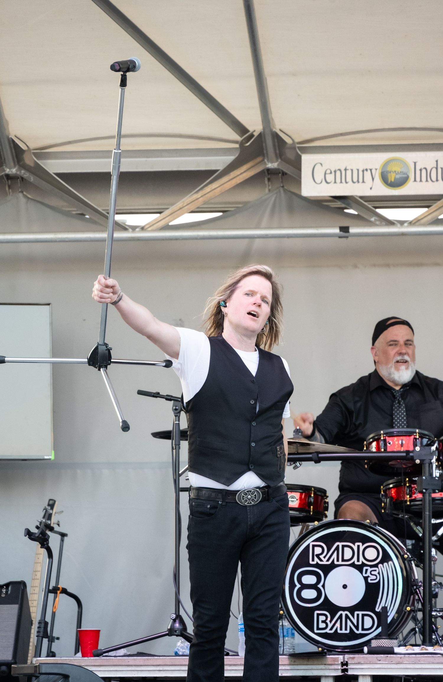 Performer singing with microphone stand on stage next to Radio 80s Band drum kit.