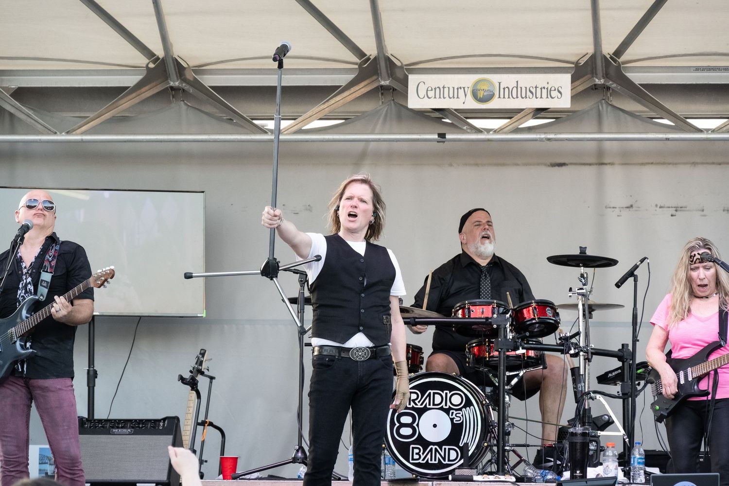 Full band performing live on outdoor stage with Radio 80s Band logo visible on drum kit.